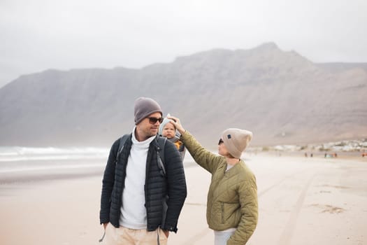 Young father carrying his infant baby boy son in backpack on windy sandy Famara beach, Lanzarote island, Spain. Family travel and winter vacation concept