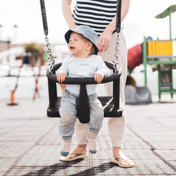 Mother pushing her infant baby boy child on a swing on playground outdoors