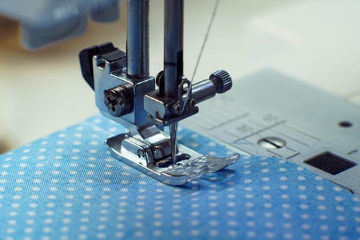 Macro shot of sewing machine steel pressure foot with needle and thread making stitches on blue fabric polka dot fabric. Tailor and dressmaker shop, atelier, dressmaker industry. Clothes making