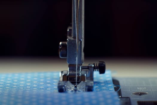 Detailed close up shot of sewing machine steel foot with needle and thread stitching on blue fabric polka dot textile. Sewing machine, tailor's shop, atelier, dressmaker industry. Needle craft