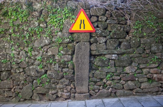 Narrowing yellow sign attached to old stone wall of rural area. Driving at villages concept