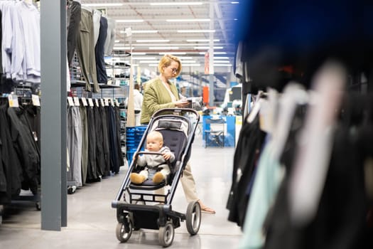 Casualy dressed mother choosing sporty shoes and clothes products in sports department of supermarket store with her infant baby boy child in stroller