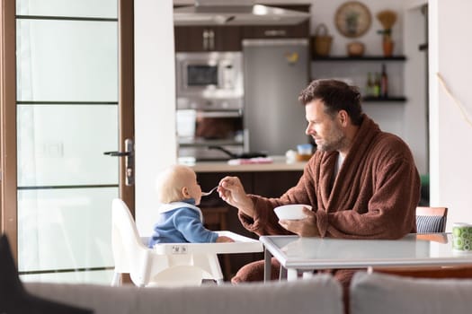 Father wearing bathrope spoon feeding hir infant baby boy child sitting in high chair at the dining table in kitchen at home in the morning