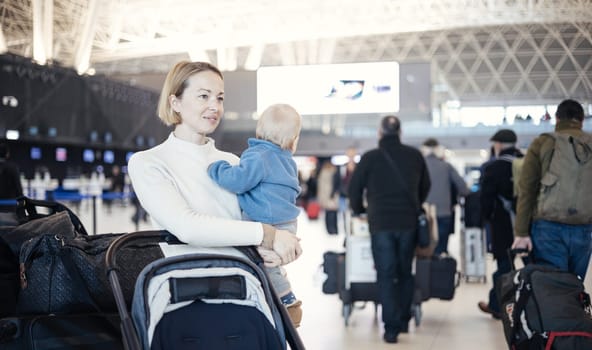 Motherat travelling with his infant baby boy child, walking, pushing baby stroller and luggage cart at airport terminal station. Travel with child concept
