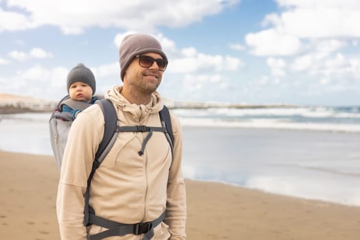 Young father carrying his infant baby boy son in backpack on windy sandy Famara beach, Lanzarote island, Spain. Family travel and winter vacation concept
