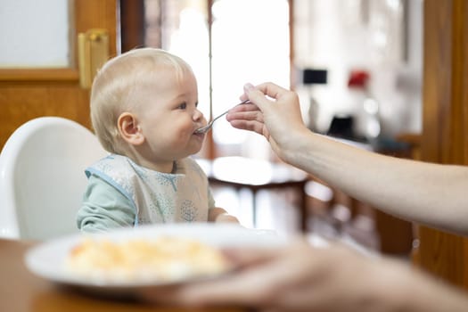 Mother spoon feeding her infant baby boy child sitting in high chair at the dining table in kitchen at home.