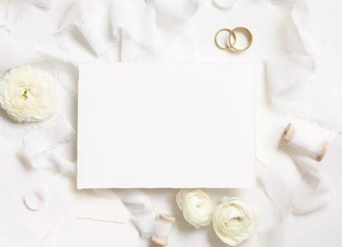 Blank card near cream roses, white silk ribbons and wedding rings top view, wedding mockup. Romantic scene with vertical card and pastel flowers flat lay. Valentines, Spring or Mothers day concept