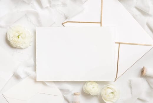 Blank card and envelope near cream roses and white silk ribbons top view, wedding mockup. Romantic scene with horizontal card and pastel flowers flat lay. Valentines, Spring or Mothers day concept