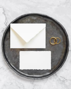 Black plate with a blank card, envelope and wto wedding rings top view, mockup. Elegant flat lay with horizontal greeting or Invitation paper card and envelope, wedding or holiday stationery