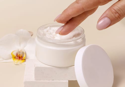 A woman hand using cream from Opened cosmetic jar with a blank lid near orchid flowers on beige close up, mockup. Skincare beauty product package, minimal composition..