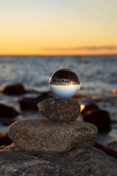 Glass ball lies on the top stone of stacked stones, at sunset on the beach, the sea and the setting sun are reflected in the ball