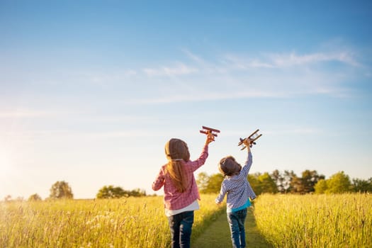Boy and girl plaing with wooden planes on the field in sunset. Concept of the family vacation, dreaming and friendship.