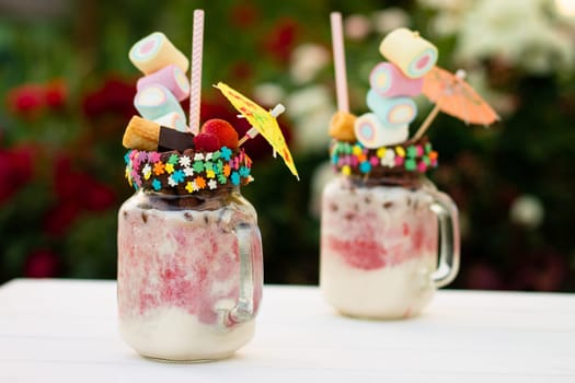 Freakshakes in jars with cocktail umbrella, straw, marshmallow, strawberry, chocolate on a white table on a background of summer flowers.