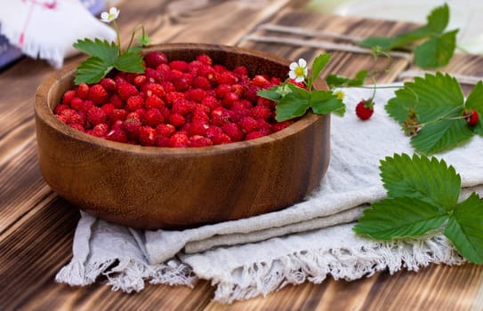 A wooden bowl of red ripe wild strawberries and flowers on an old wooden surface.