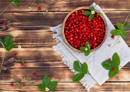 A wooden bowl of red ripe wild strawberries and flowers on an old wooden surface. Top view. Copy space.