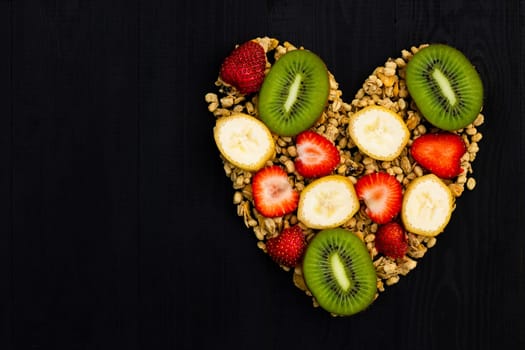 Heart laid out of granola and fruits on a black wooden background. Flat lay. Copy space.