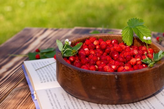 A wooden bowl of red ripe wild strawberries and flowers with a book on an old wooden surface. Copy space.