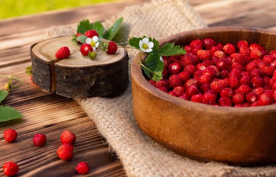 A wooden bowl of red ripe wild strawberries and flowers on an old wooden surface.