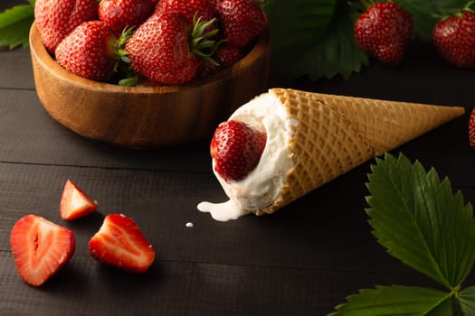 Ice cream and wooden bowl of fresh red ripe strawberries on a black wooden surface. Low key.