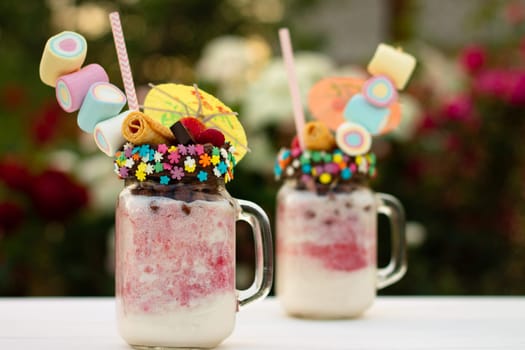 Freakshakes in jars with cocktail umbrella, straw, marshmallow, strawberry, chocolate on a white table on a background of summer flowers.