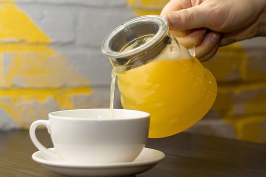 Hand pours yellow ginger tea with lemon and rosemary branch from transparent teapot into white cup on dark wooden table