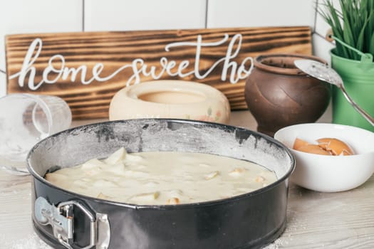 Baking dish with raw apple pie dough stands on kitchen counter next to ingredients, cooking utencils and wooden sign Home sweet home