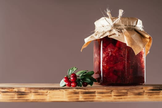Jar of homemade lingonberry and pear jam with craft paper on lid on wooden shelf next to fresh lingonberries on brown background