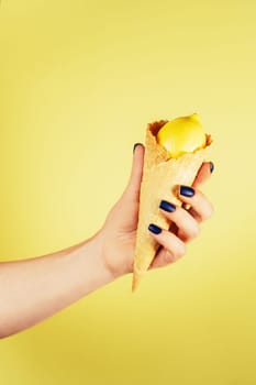 Female hand with blue nails holding waffle ice cream cone filled with fresh ripe juicy lemon isolated on yellow. Creative art concept. Food minimal colorful concept. Vertical. Copy space. Mock up.