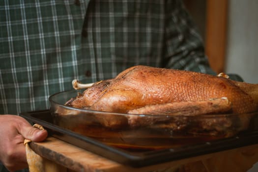 Man in green checkered shirt carries baked goose or turkey, on baking sheet
