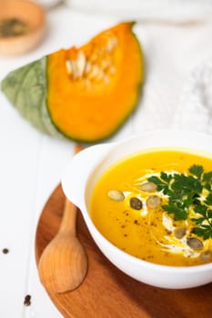 A plate of yellow pumpkin cream soup with cream, herbs and seeds on a white table near ginger, raw pumpkin and a wooden spoon. Vertical