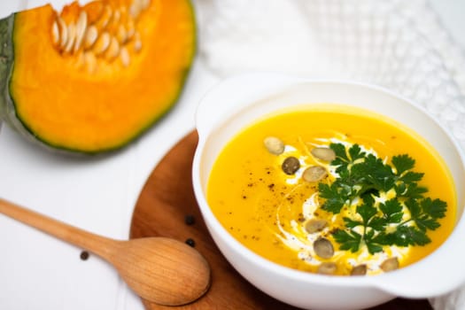 A plate of yellow pumpkin cream soup with cream, herbs and seeds on a white table near ginger, raw pumpkin and a wooden spoon.