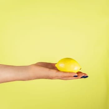Female hand with blue nails holding fresh ripe juicy lemon isolated on yellow. Squeeze out juice. Diet topic. Creative art concept. Food minimal colorful concept. Vertical. Copy space. Mock up.