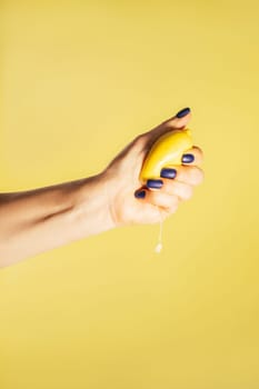 Female hand with blue nails holding tight fresh ripe juicy lemon isolated on yellow. Squeeze out juice. Diet topic. Creative art concept. Food minimal colorful concept. Vertical. Copy space. Mock up.