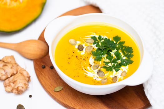 A plate of yellow pumpkin cream soup with cream, herbs and seeds on a white table near ginger, raw pumpkin and a wooden spoon.