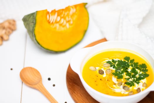 A plate of yellow pumpkin cream soup with cream, herbs and seeds on a white table near ginger, raw pumpkin and a wooden spoon.