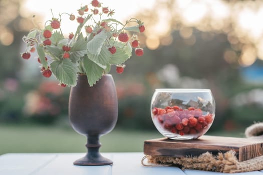 Fresh ripe strawberry bush with green leaves in clay brown vase in shape of glass and peeled strawberries in glass bowl on wooden board on woolen brown cloth. Organic background. Selective focus.