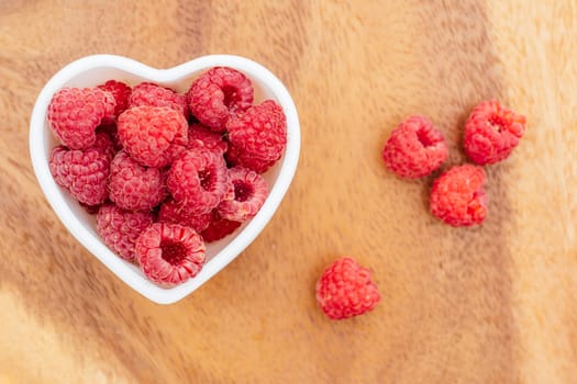 Heart shaped bowl full of ripe red juicy raspberries on wooden background. Top view. Copy space