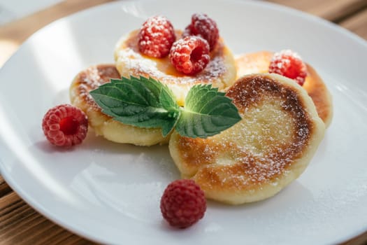 Delicious cheesecakes or pancakes sprinkled with powdered sugar on white plate on wooden table with raspberries and mint leaf