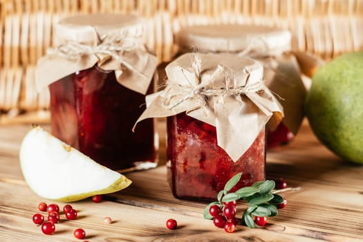 Jars of homemade lingonberry and pear jam with craft paper on lids on wooden surface next to fresh lingonberries and pears.