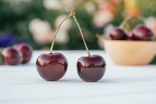 Two red ripe sweet cherries berries standing on wooden table on background of multi-colored blurred bushes with flowers next to bowl of sweet cherries