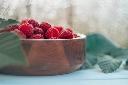 Partially blurred wooden round bowl full of ripe red juicy raspberries on white wooden surface against bokeh background with raspberry bush leaves