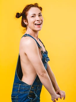Sexy topless redhead woman laughing and wearing overalls over yellow background.