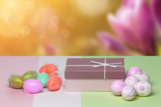 Easter greeting card template. Close-up of colorful eggs and a gift box on a multicolor table against abstract spring background. Copy space for your text design.