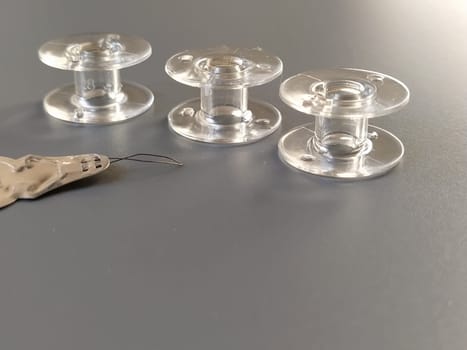 Buttonhole for automatically threading the sewing machine needle. Three transparent plastic sewing machine bobbins. Textile industry. Detail of sewing equipment. Gray background, back lighting.