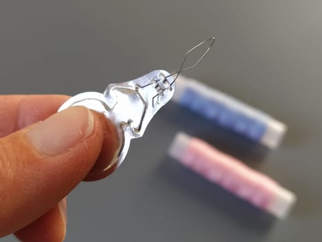 device for drawing the thread into the eye of the needle. Bobbins with blue and pink threads in the background. A woman holds a needle threader in her hand and shows its thin wire eye.