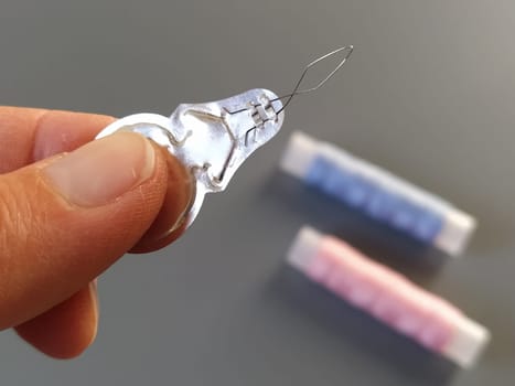 device for drawing the thread into the eye of the needle. Bobbins with blue and pink threads in the background. A woman holds a needle threader in her hand and shows its thin wire eye.
