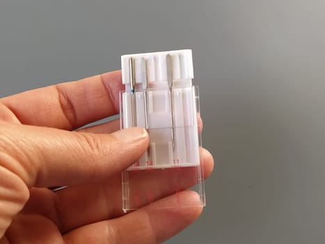 a set of sewing machine needles in a white plastic case. A woman's hand holds the needles for an electric sewing machine. Gray background. Different needle thicknesses.