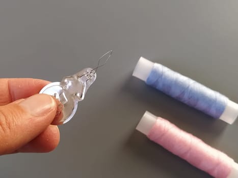device for drawing the thread into the eye of the needle. Bobbins with blue and pink threads in the background. A woman holds a needle threader in her hand and shows its thin wire eye.