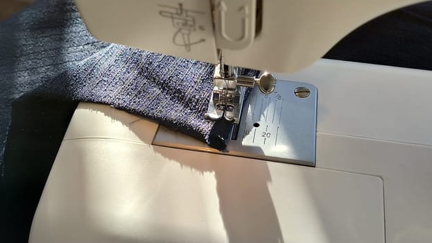 Sewing process on a modern sewing machine, close-up. Sewing machine foot with a needle. Dark blue fabric is trimmed with double wrapping of the fabric edge. Natural sunlight.