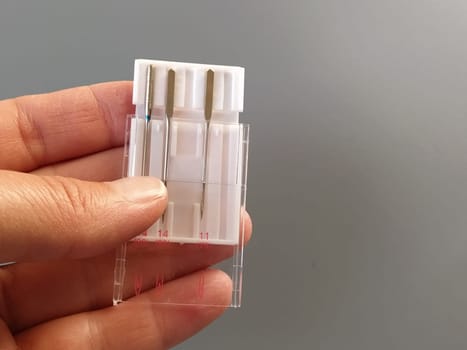 a set of sewing machine needles in a white plastic case. A woman's hand holds the needles for an electric sewing machine. Gray background. Different needle thicknesses.
